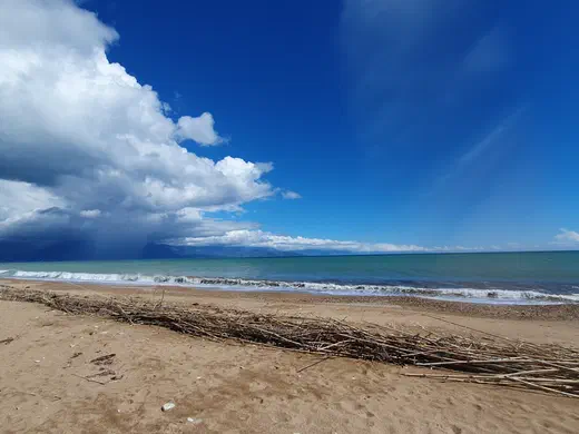 Sandstrand mit Schilf, dahinter das türkisfarbene Meer und eine Regenwolke