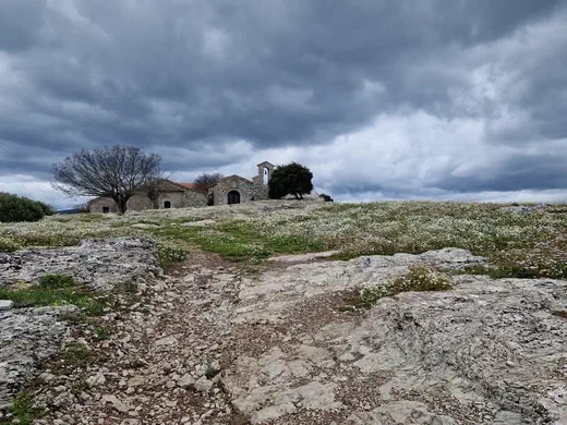 Steinkapelle auf einem Hügel unter dunklen Wolken, ringsum eine blühende Wiese