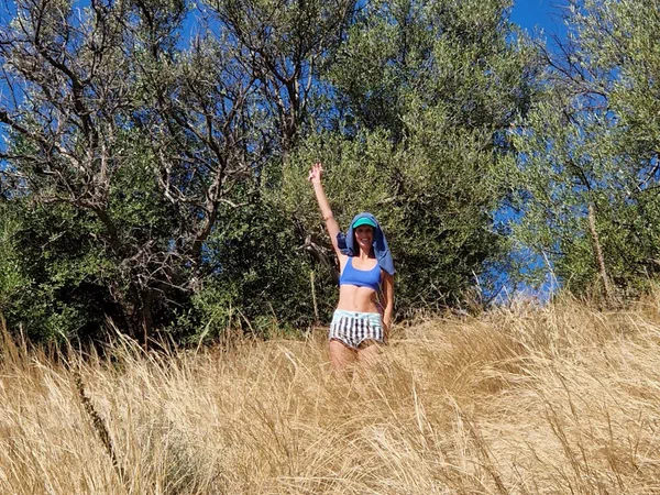 Stefanie stands with a raised arm in tall dry grass in front of an olive tree