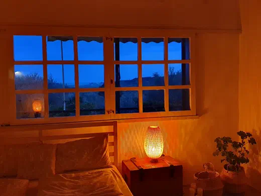 Schlafzimmer im warmen Lampenlicht, durch das Sprossenfenster der blaue Abendhimmel
