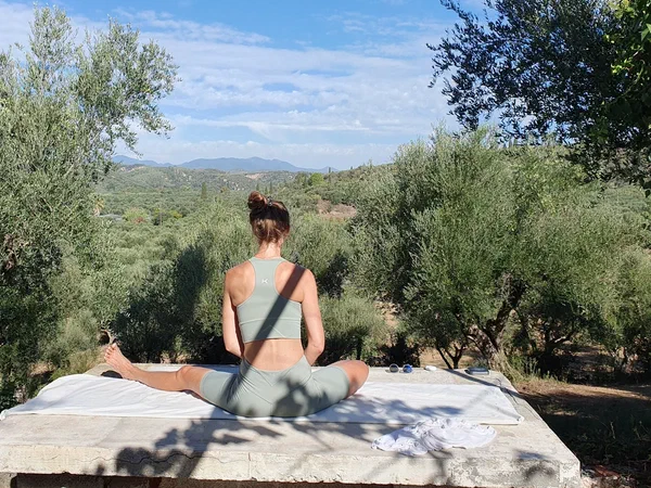A woman sits in a yoga posture on a stone platform looking out across the olive groves