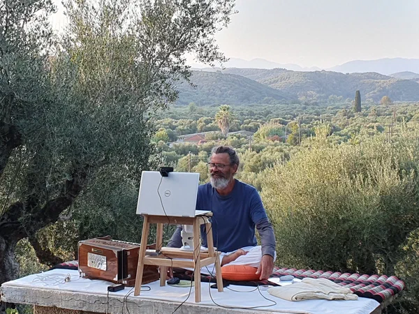 Clemens sits with a laptop and a harmonium on the terrace, olive groves and mountains behind him