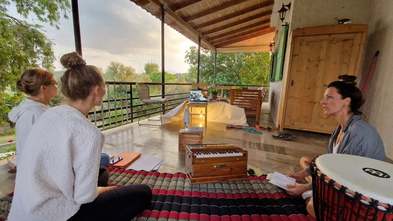 Three people sit talking on mats on a covered terrace, with a harmonium and a drum beside them