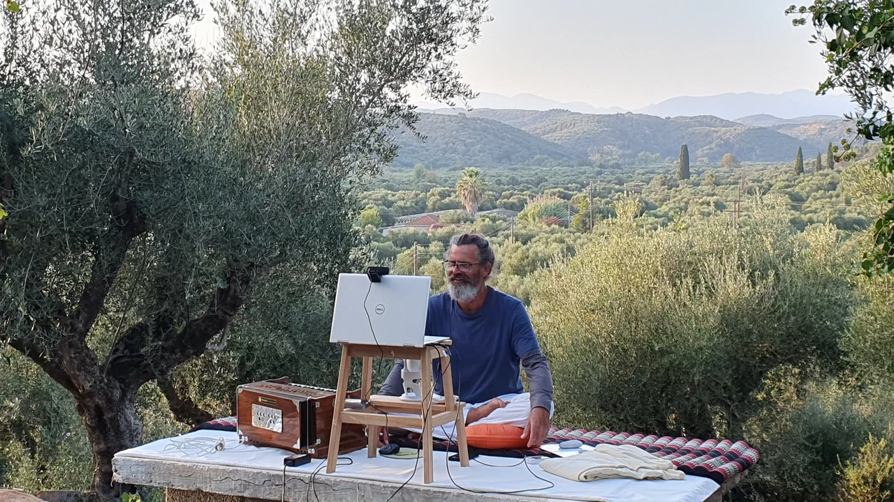 Clemens sits with a laptop and a harmonium on the terrace, olive groves and mountains behind him