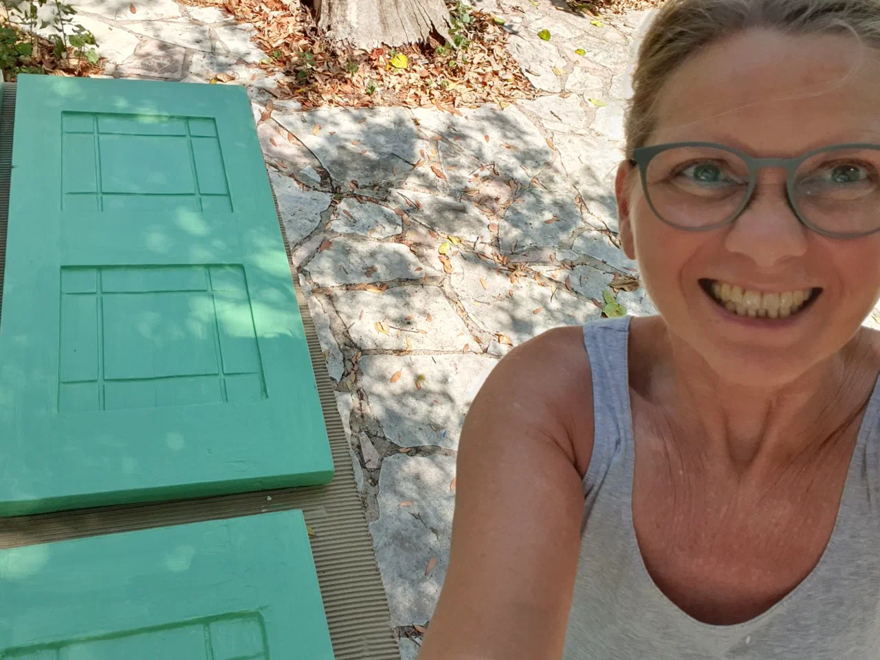 Stefanie painting window shutters turquoise, in the shade of a tree