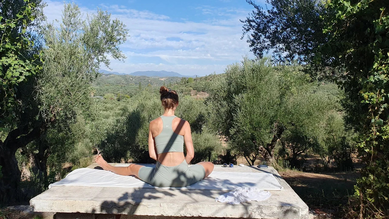 A woman sits in a yoga posture on a stone platform, looking out across the olive groves towards the mountains